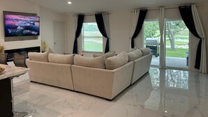 Un salon moderne avec un canapé d’angle beige, un téléviseur à écran plat fixé au mur et une grande fenêtre avec des rideaux.