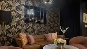 Una habitación con papel tapiz estampado, un sofá de cuero marrón con cojines, una mesa redonda de mármol con un jarrón de flores y una lámpara de pie.
