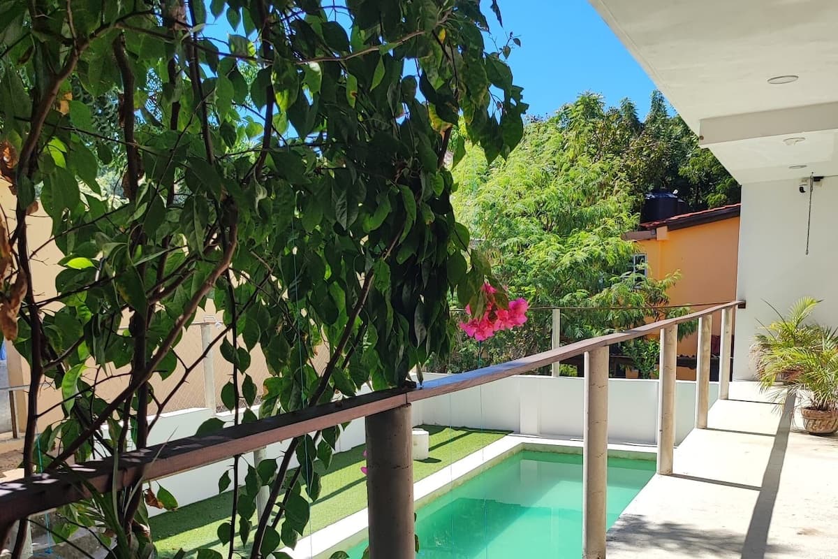 A pool area with a clear view of the pool, surrounded by greenery and a white building.