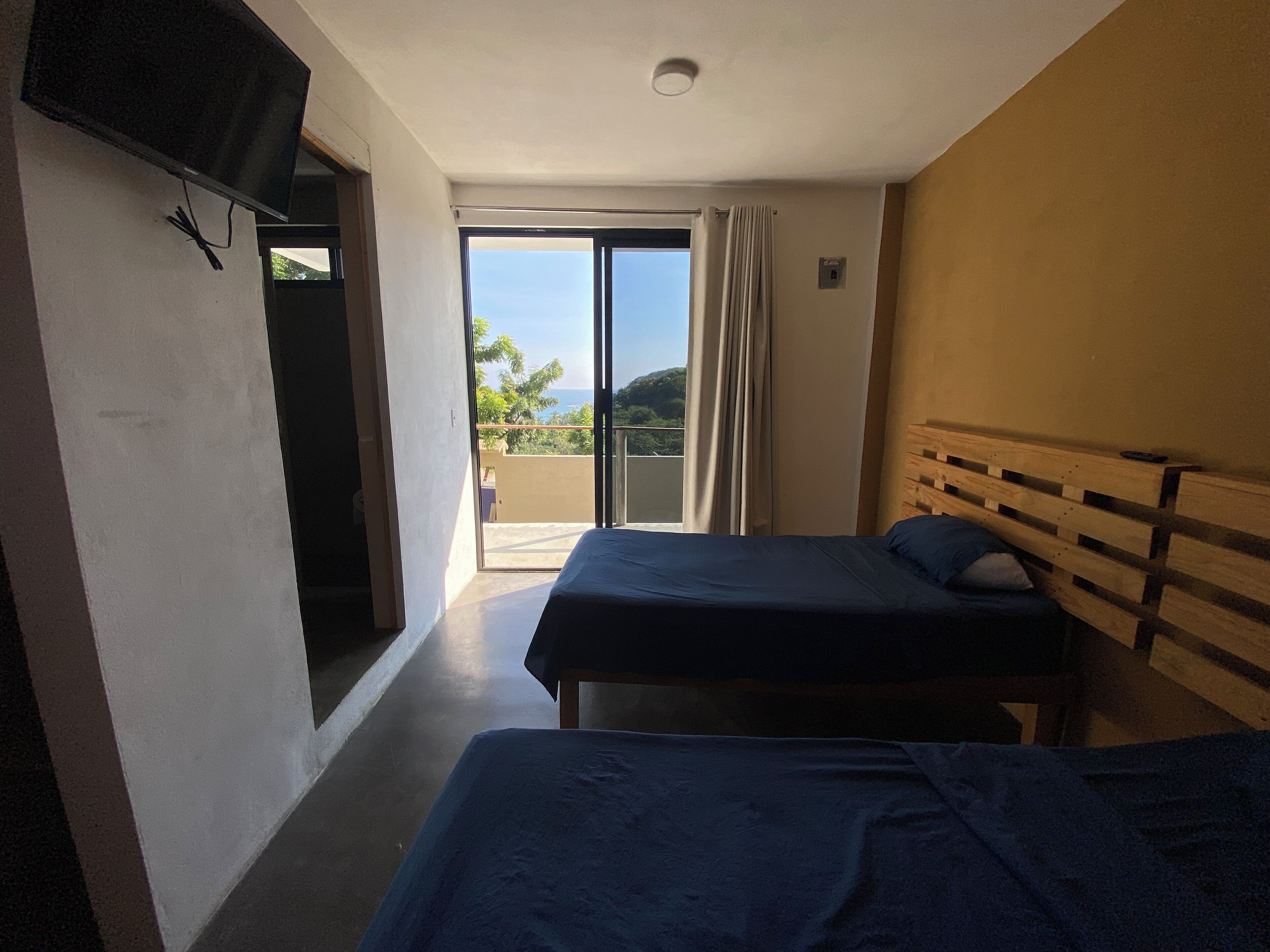 Ein Hotelzimmer mit zwei Betten, einem an der Wand montierten Fernseher und einem Balkon mit Blick auf Grünflächen und Hügel.
