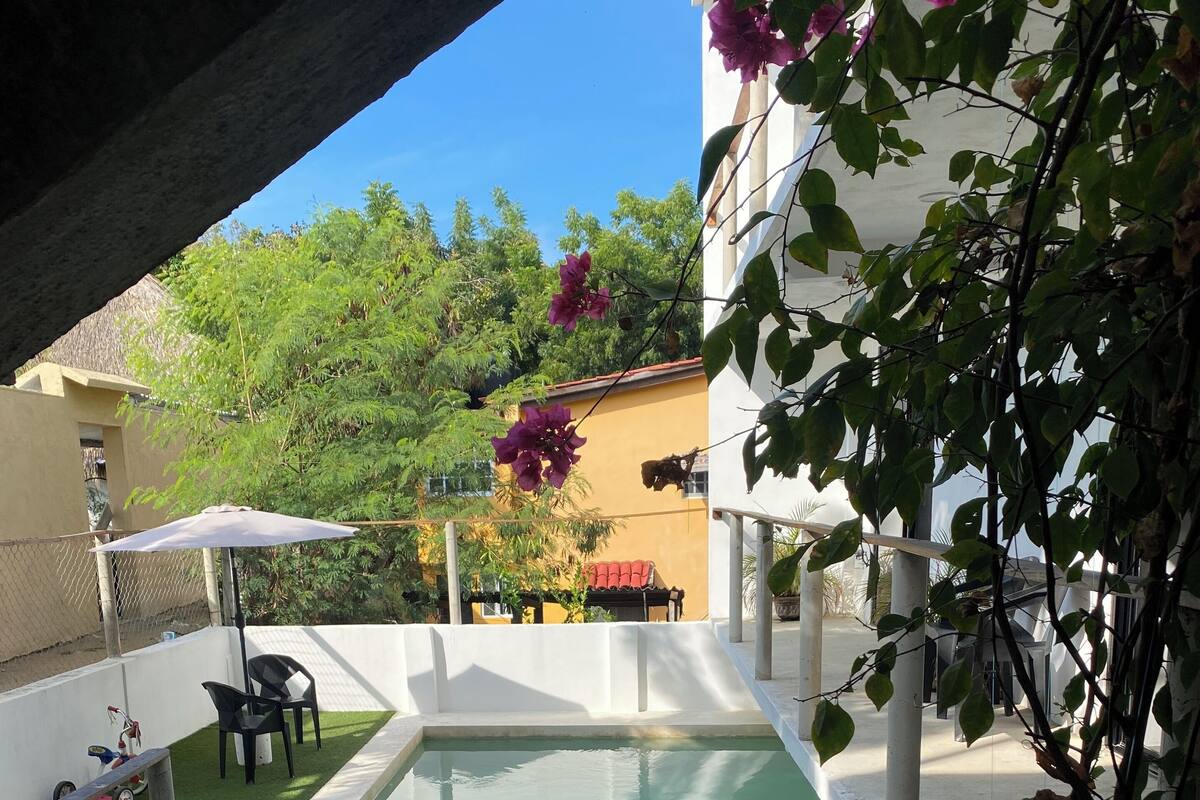 A swimming pool with a small deck area, surrounded by a fence, and a chair under an umbrella.