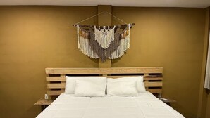 Una habitación de hotel con cabecera de madera, cama con ropa de cama blanca y un tapiz de macramé en la pared.