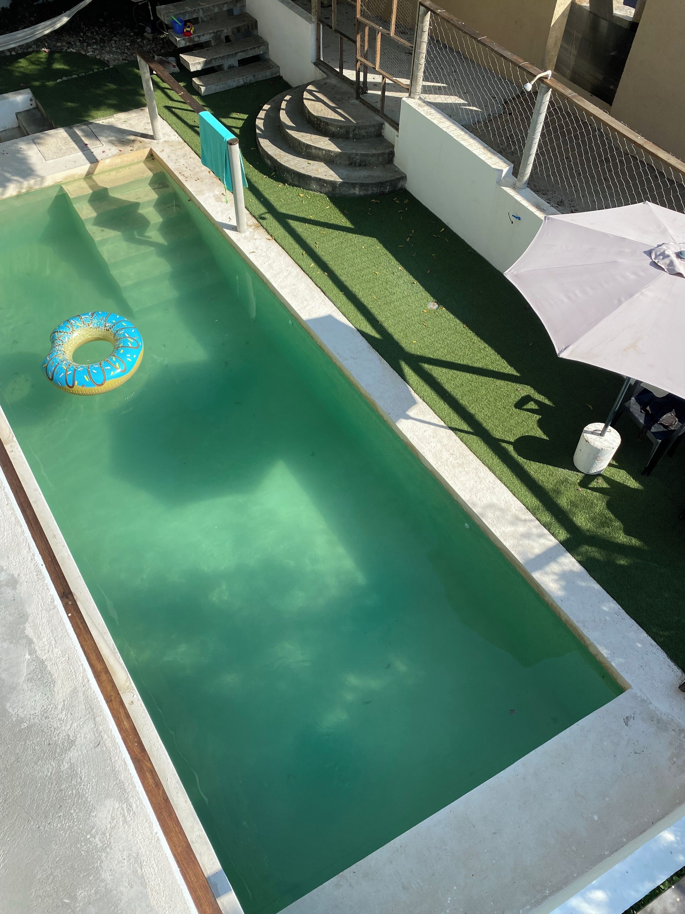 Une piscine avec un flotteur bleu gonflable, des marches menant à une terrasse et un parasol blanc.