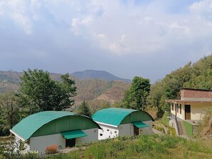 A rural setting with two green-roofed structures, one with a visible door and windows, and a staircase leading to a building with a red roof. - Oak Retreat Huts (Shimla)