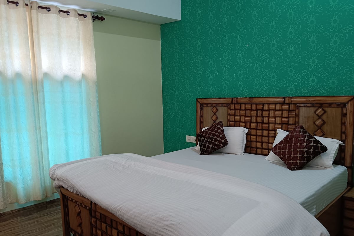 A neatly made bed with a wooden headboard, a green patterned wall, and a window with a white curtain.
