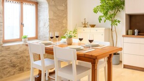 Un coin repas avec une table en bois dressée pour quatre personnes, des chaises blanches, une plante en pot et une fenêtre avec des rideaux.
