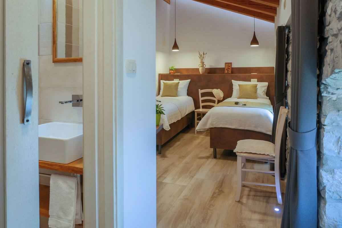 Een slaapkamer met twee bedden, een houten hoofdbord, een nachtkastje, een stoel, een hanglamp en een badkamer met een wastafel en handdoekhouder.