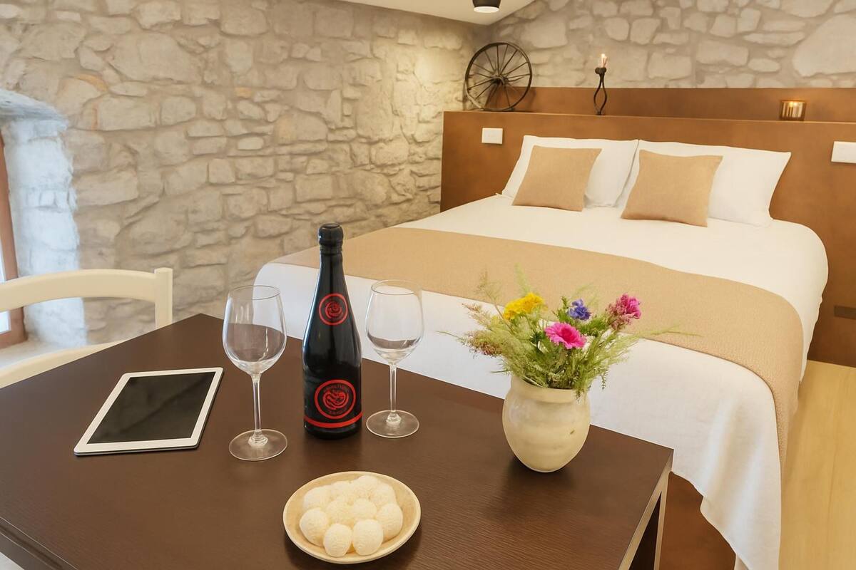 Hotelkamer met een stenen muur, een houten hoofdbord, een bed met wit en beige beddengoed, een tafel met een tablet, wijnglazen, een fles en een vaas met bloemen.