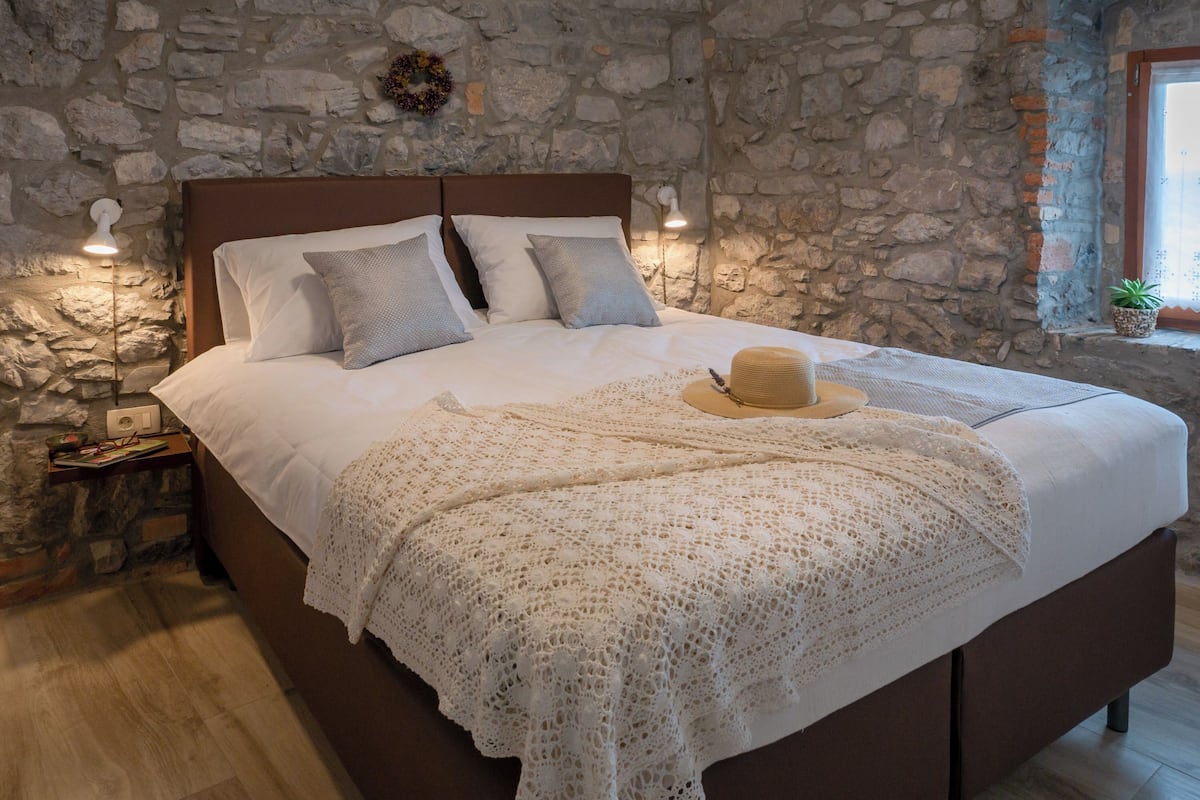 Een slaapkamer met een stenen muur, een houten hoofdbord en een bed met witte lakens en een witte kantdeken.