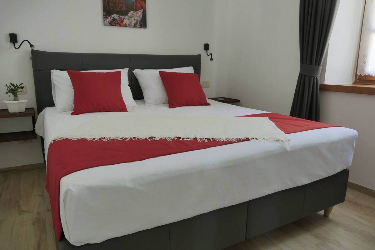 Een netjes opgemaakt bed met wit en rood beddengoed, een donker hoofdbord en een klein nachtkastje met een plantje.