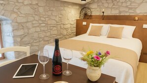 Habitación de hotel con pared de piedra, cabecera de madera, cama con ropa de cama blanca y beige, una mesa con una tableta, copas de vino, una botella y un jarrón con flores.