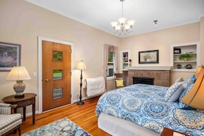 Quarto com uma cama grande, porta de madeira, abajur, cadeira, lareira e estante com objetos decorativos.