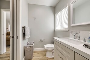 A bathroom with a toilet, sink, and mirror. There is a trash can and a towel hanging on the wall.