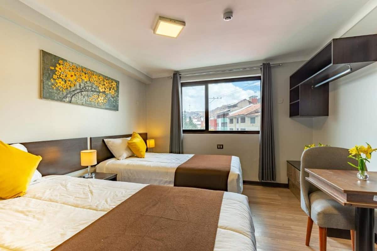 Quarto de hotel com duas camas, uma escrivaninha, uma cadeira, um quadro na parede e uma janela com vista para a cidade.