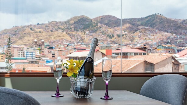 A table with a bottle of champagne, two glasses, and a vase with flowers, set against a cityscape view. - Tikala Cusco Boutique Hotel (Cusco)