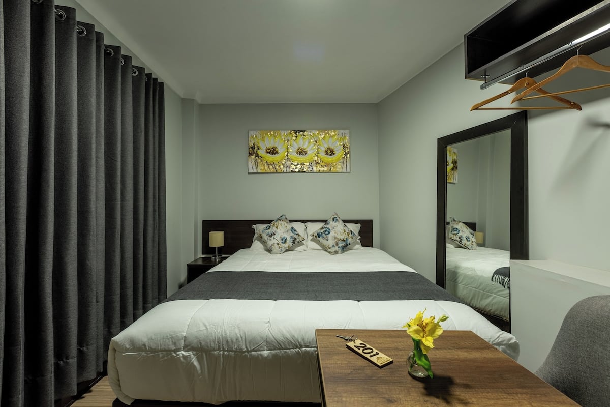 Quarto de hotel com uma cama grande, mesa de cabeceira de madeira, espelho, obra de arte na parede e escrivaninha com um vaso de flores.