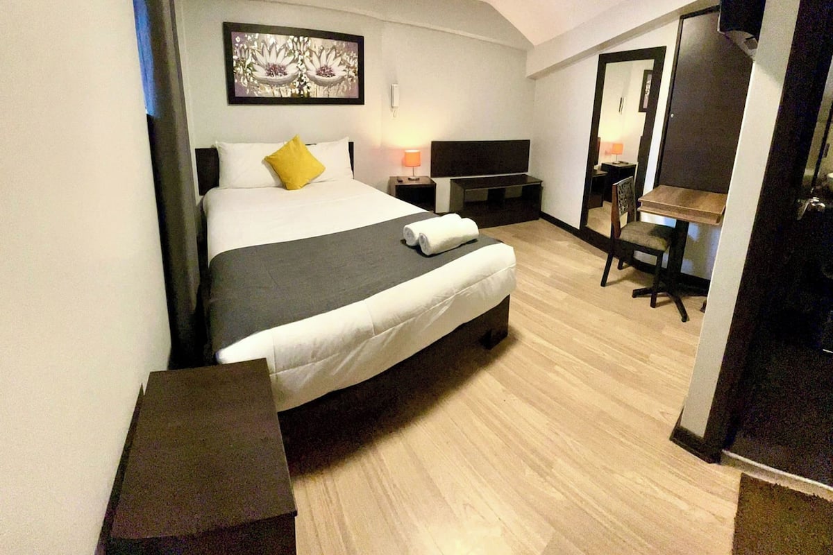 Quarto de hotel com cama, mesa de cabeceira, televisão e escrivaninha com cadeira.