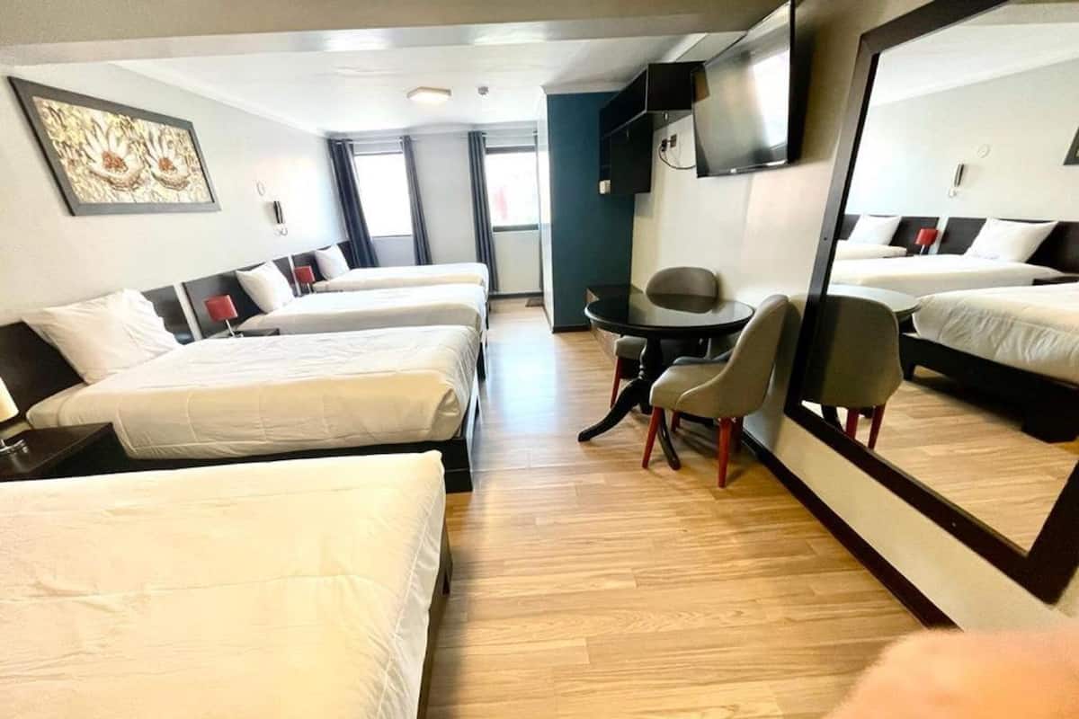 Quarto de hotel com três camas de solteiro, uma mesa redonda pequena com duas cadeiras, um espelho grande e uma TV fixada na parede.