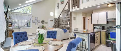 Amplia sala de estar con mesa de comedor, sillas tapizadas en azul, cocina con electrodomésticos modernos y una escalera que conduce a un nivel superior.