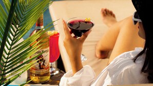 Une personne se détend près d’une piscine, assise sur une chaise longue, avec un verre à la main et une bouteille d’alcool posée sur une table.