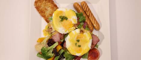 Une assiette carrée blanche contenant un petit-déjeuner composé d’œufs, de bacon, de saucisses et de légumes.
