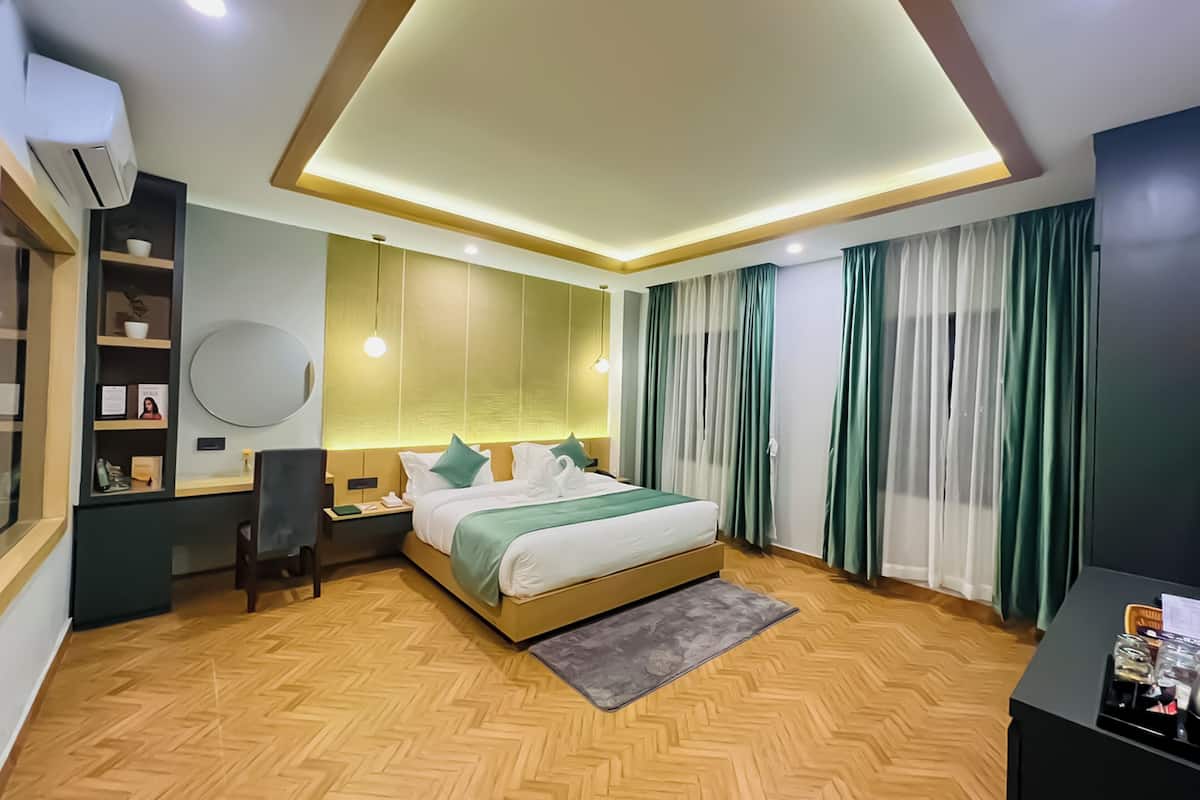A modern hotel room with a wooden herringbone floor, a bed with a green and white bedspread, a desk with a chair, a round mirror, and a nightstand.
