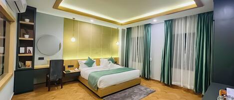 A modern hotel room with a wooden herringbone floor, a bed with a green and white bedspread, a desk with a chair, a round mirror, and a nightstand.