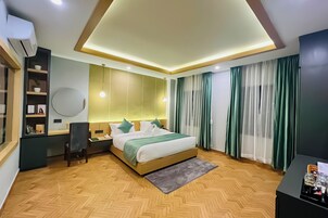 Una habitación de hotel moderna con piso de madera en patrón de espiga, cama con colcha verde y blanca, escritorio con silla, espejo redondo y mesita de noche.