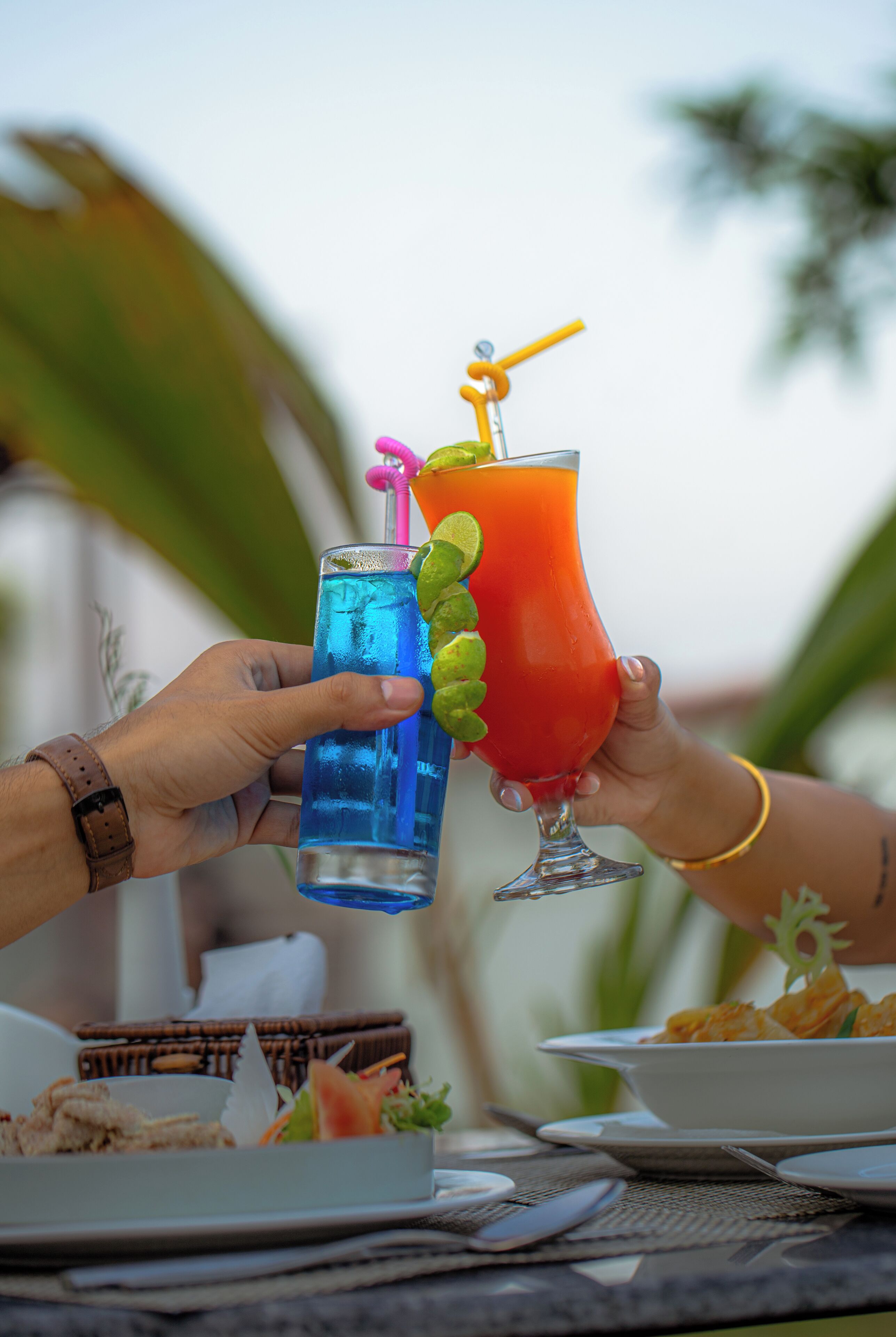 Dos personas sosteniendo bebidas en una mesa, con un plato de comida y una cesta de mimbre al fondo.