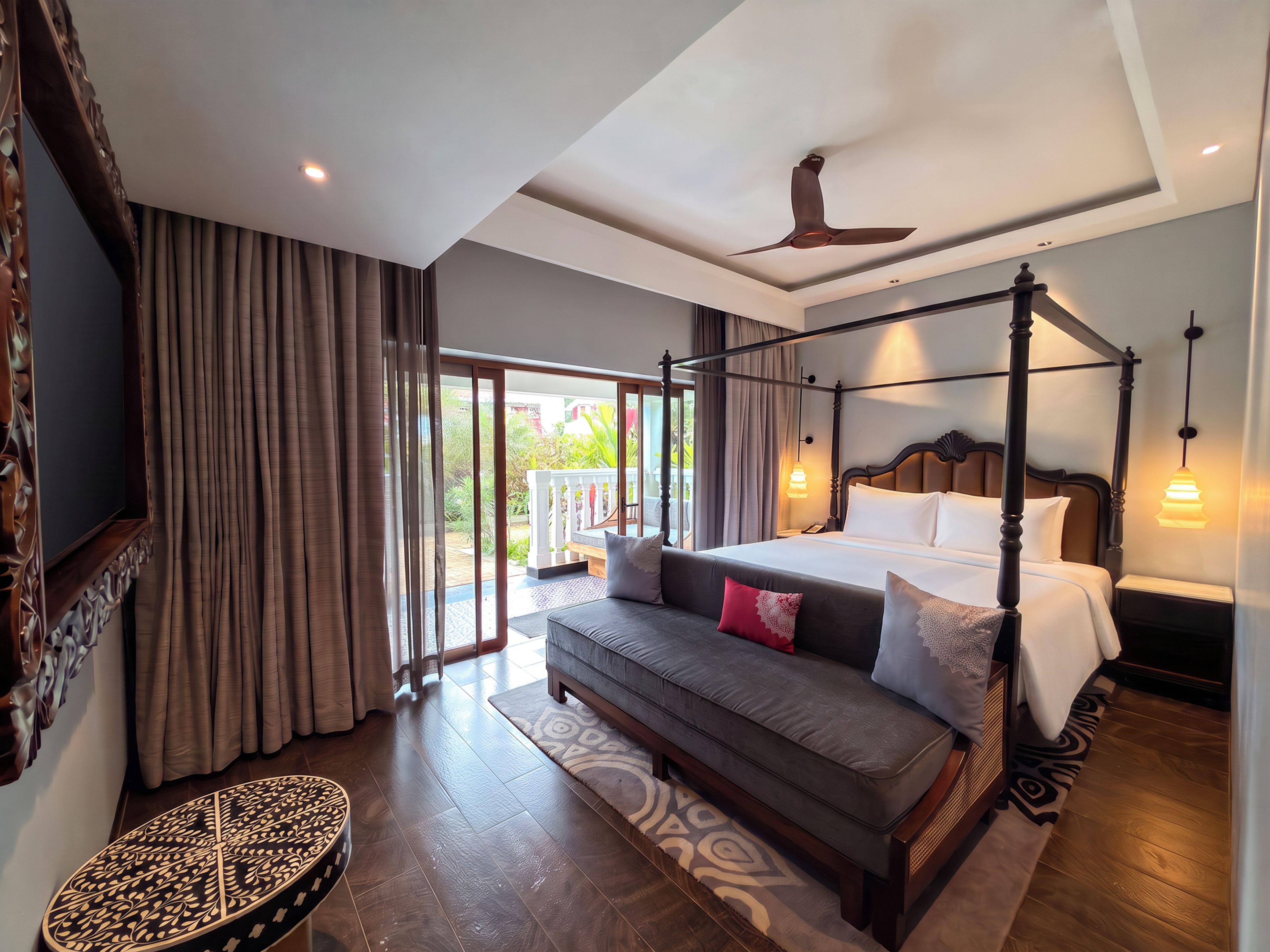 A four-poster bed with a ceiling fan, a sofa with pillows, a patterned rug, and a round table with a decorative top.