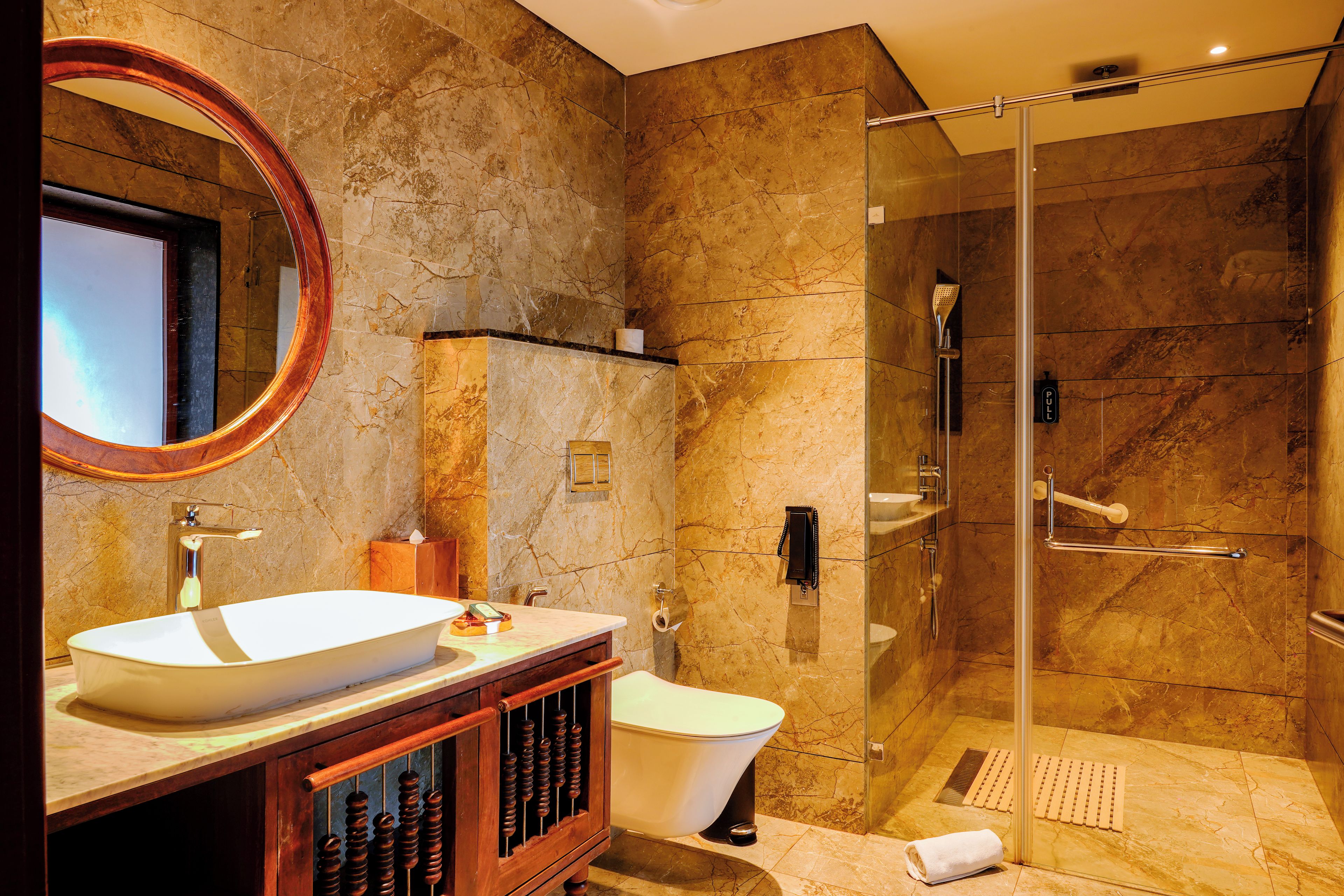 A bathroom with a marble wall, a round mirror, a white sink, a toilet, and a glass-enclosed shower area.