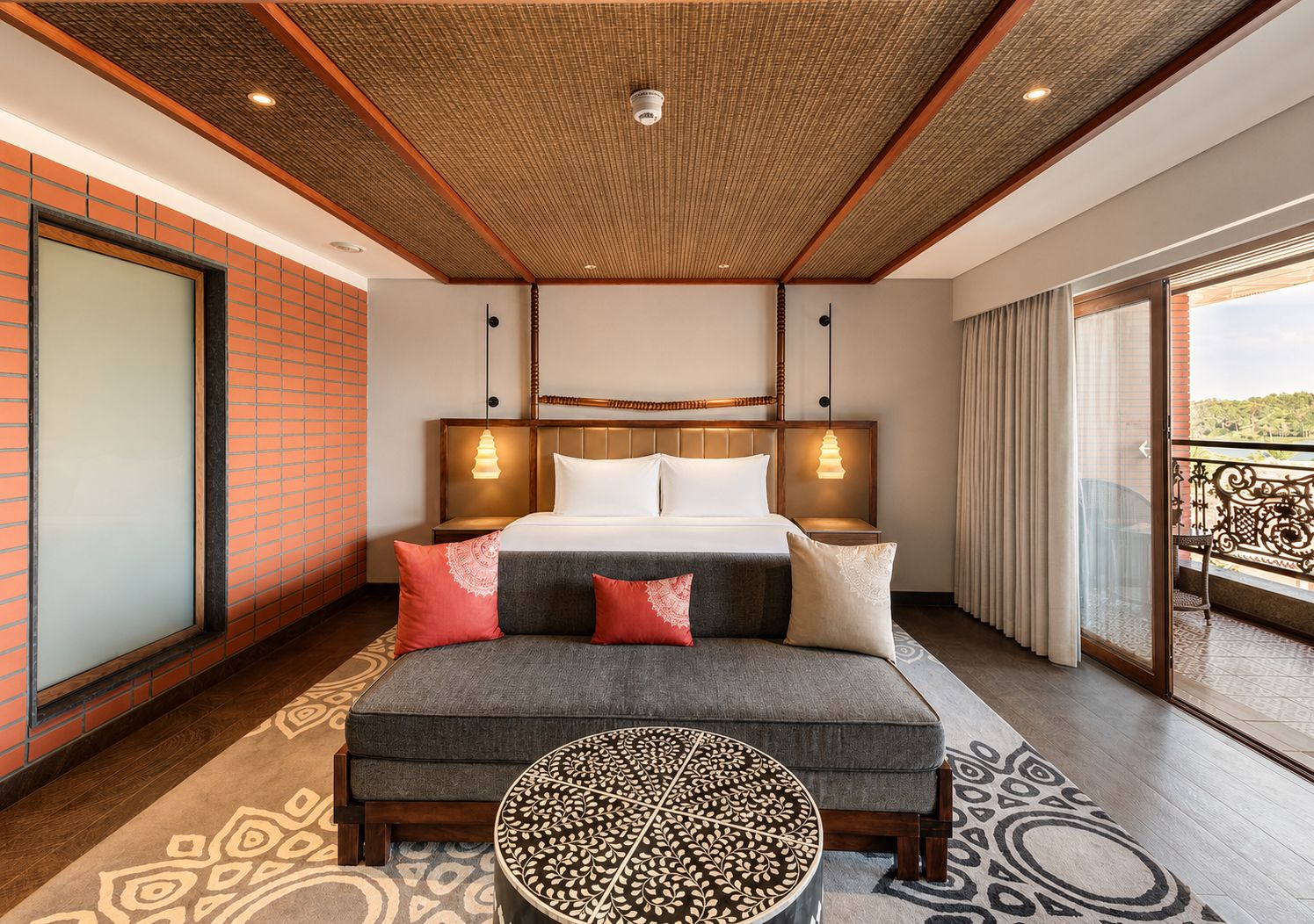 Habitación de hotel con cama, mesitas de noche, sofá, otomana estampada y balcón con barandilla.