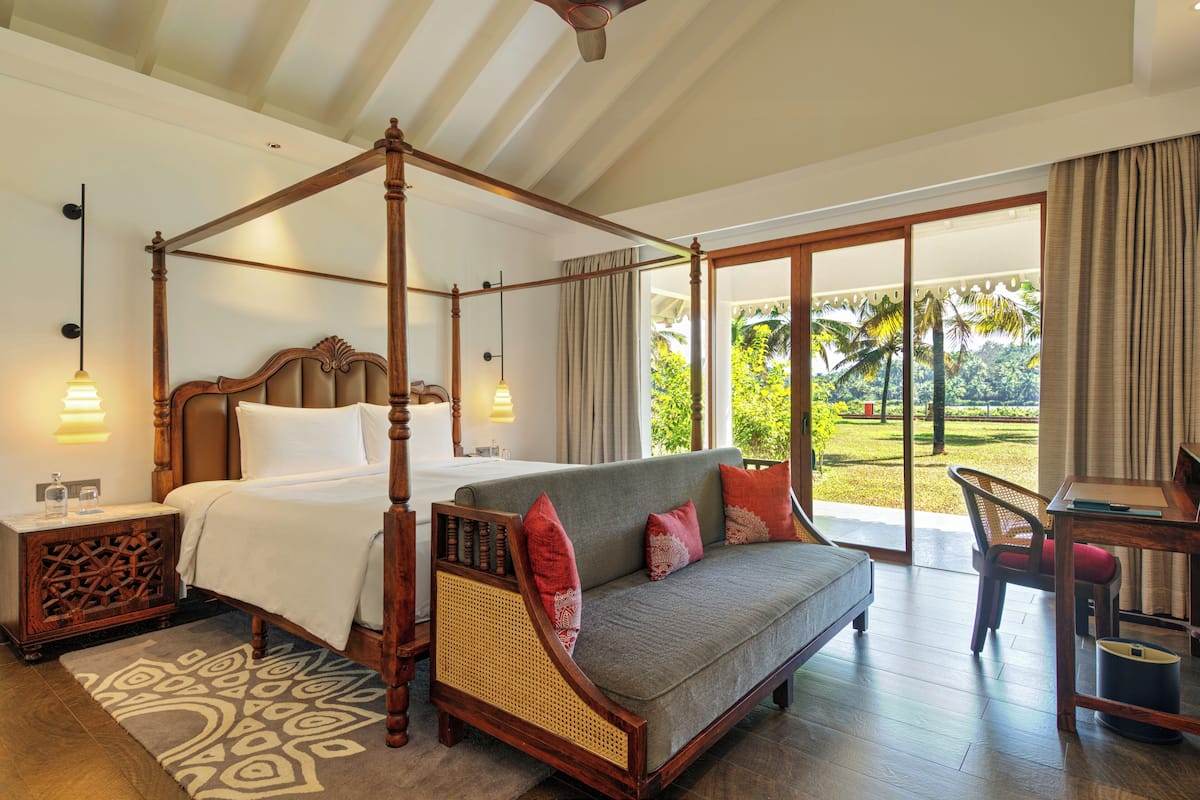 A four-poster bed with a canopy, a sofa with cushions, a dining table, and a view of a garden through a glass door.