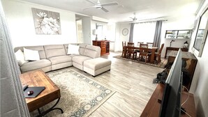 Uma sala de estar com sofá seccional, mesa de centro, mesa de jantar com cadeiras, televisão e ventiladores de teto.