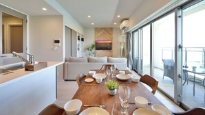 A modern dining area with a wooden table set for a meal, a sofa in the background, and a view of the outdoors through large sliding doors. - Branchera naha akebono premist R65 (Naha)