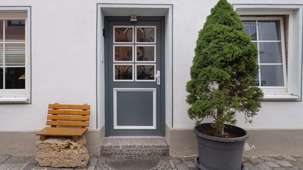 Une maison avec une porte grise, un banc en bois et un arbre en pot.
