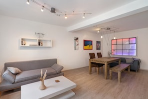 A modern living room with a grey sofa, a white coffee table, a wooden dining table with chairs, and a large window with a view.