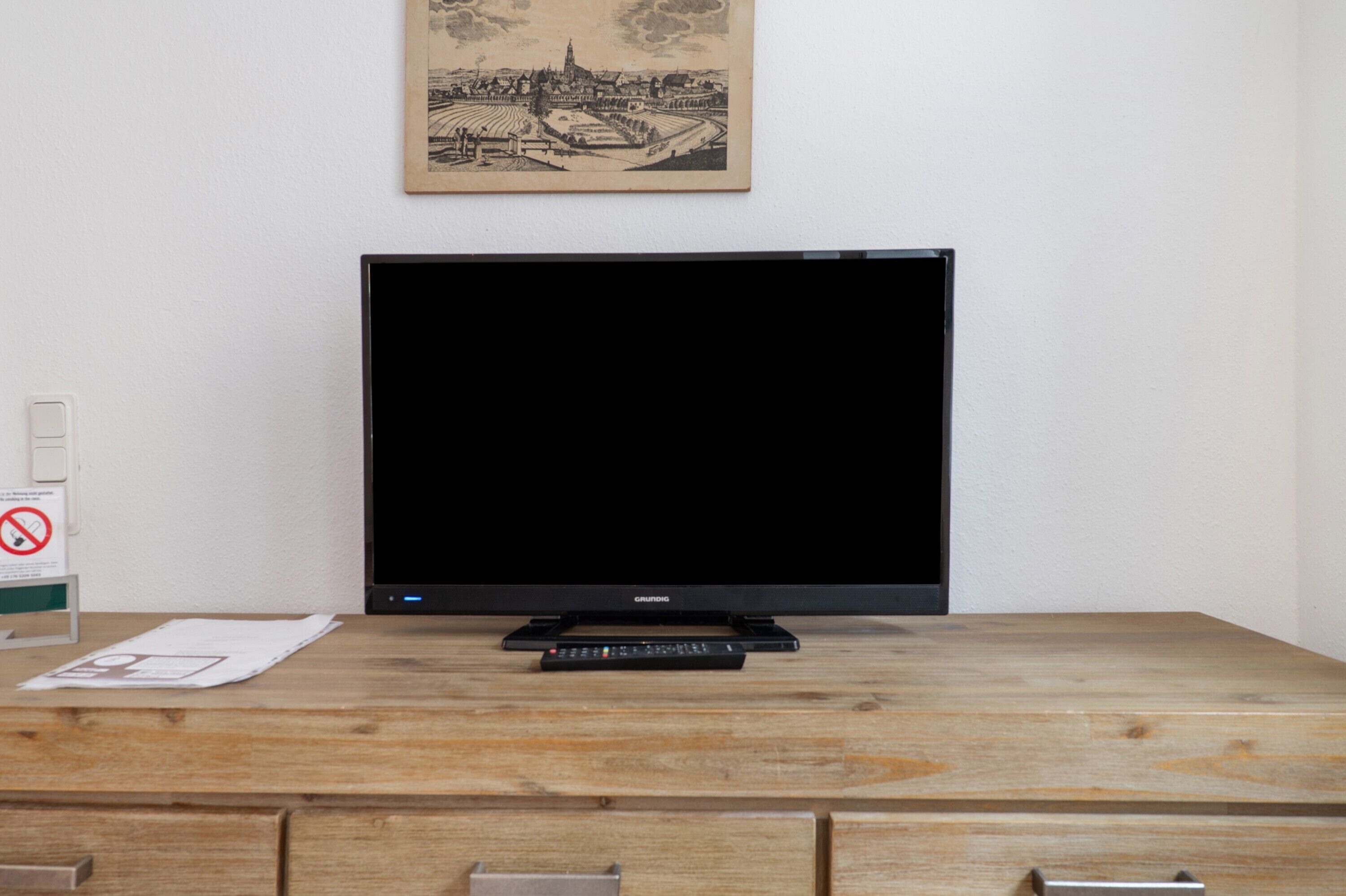 Ein Fernseher auf einem Holztisch mit Fernbedienung, ein gerahmtes Bild an der Wand und ein Rauchverbot-Schild.