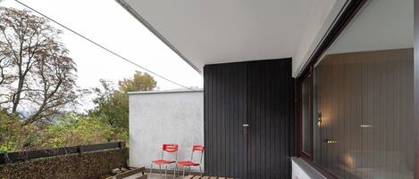 Um pátio externo com mesa e bancos de madeira, duas cadeiras vermelhas e uma janela grande.