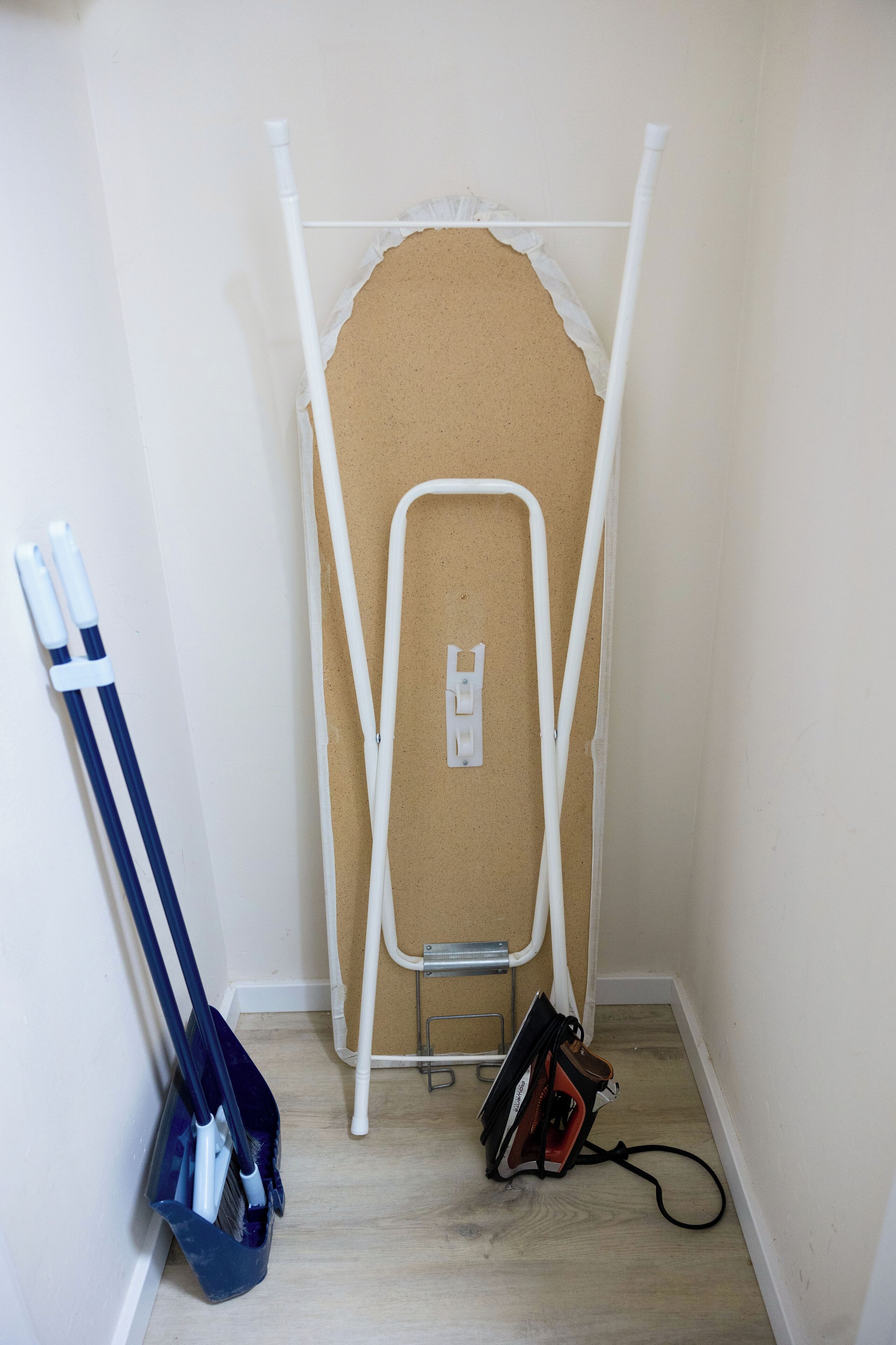 Esquina de una habitación con una tabla de planchar blanca, una escoba y un bulto en el suelo.