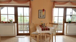 A dining area with a table set for four, wooden chairs, a white tablecloth, and a view through large windows.