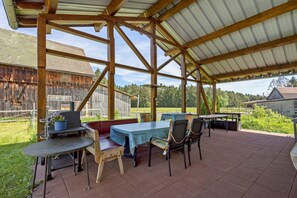Une terrasse extérieure couverte, dotée de tables et de chaises, d’un barbecue et offrant une vue sur une grange en bois et des arbres.