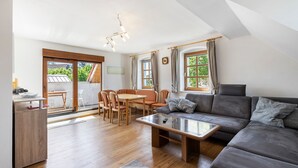 Ein modernes Wohnzimmer mit Sofa, Couchtisch, Esstisch und Stühlen. Große Fenster mit Vorhängen, eine Deckenleuchte und ein Blick ins Freie.