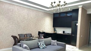 Ein modernes Wohnzimmer mit grauem Sofa, einem Esstisch mit Stühlen, einer Kristallleuchtre, und einer Küchenzeile mit dunklen Schränken und einem Kühlschrank.