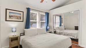 A bedroom with a large bed, a nightstand, a ceiling fan, a window with curtains, and a mirror.
