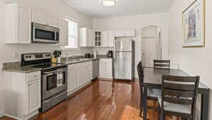 A kitchen with white cabinets, a stainless steel oven, a microwave, and a refrigerator. There is a dining table with chairs and a window with blinds.