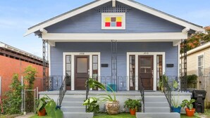 A two-story house with a blue exterior, numbered 3008 and 3010, featuring a front porch with steps, potted plants, and a small garden area.