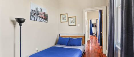 A bedroom with a bed, a ceiling fan, a floor lamp, and a cityscape painting on the wall.