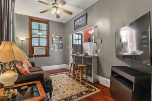 A living room with a sofa, a coffee table, a lamp, a ceiling fan, a TV, a kitchen area with a fridge, and a window with blinds.