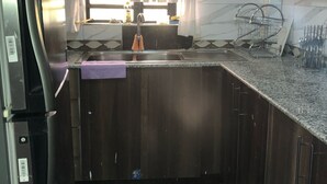 A compact kitchen with dark wooden cabinets, a stainless steel refrigerator, a marble backsplash, and a window with sheer curtains.